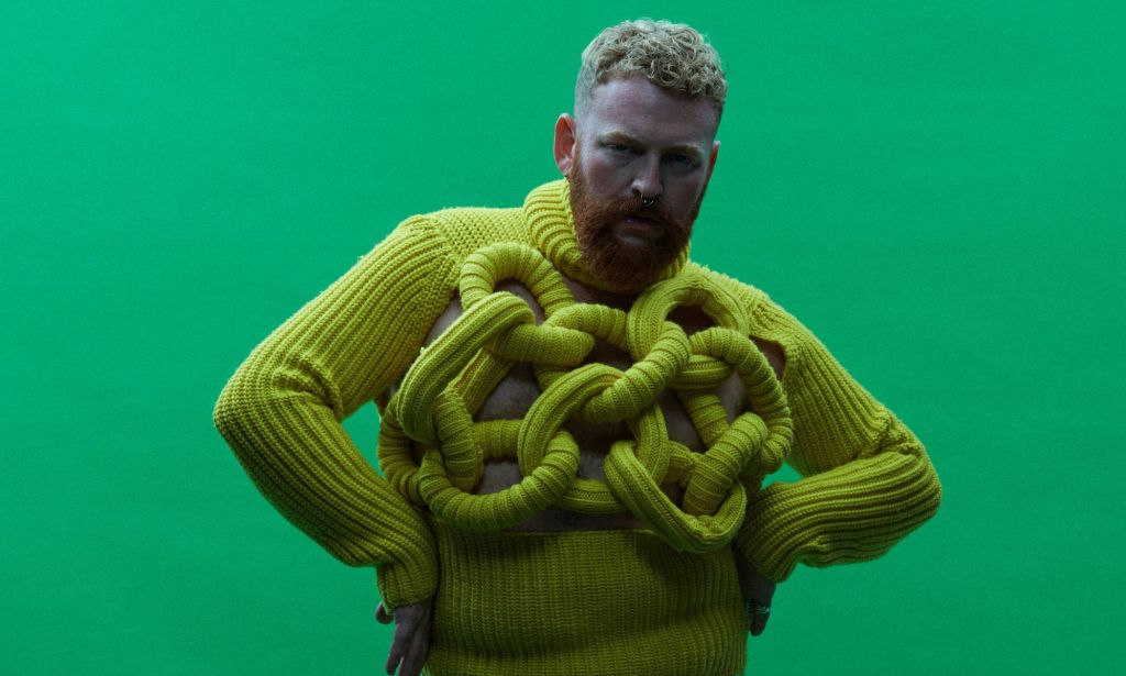 Non-binary, trans singer Tom Rasmussen wears a yellow sweater while standing in front of a green background