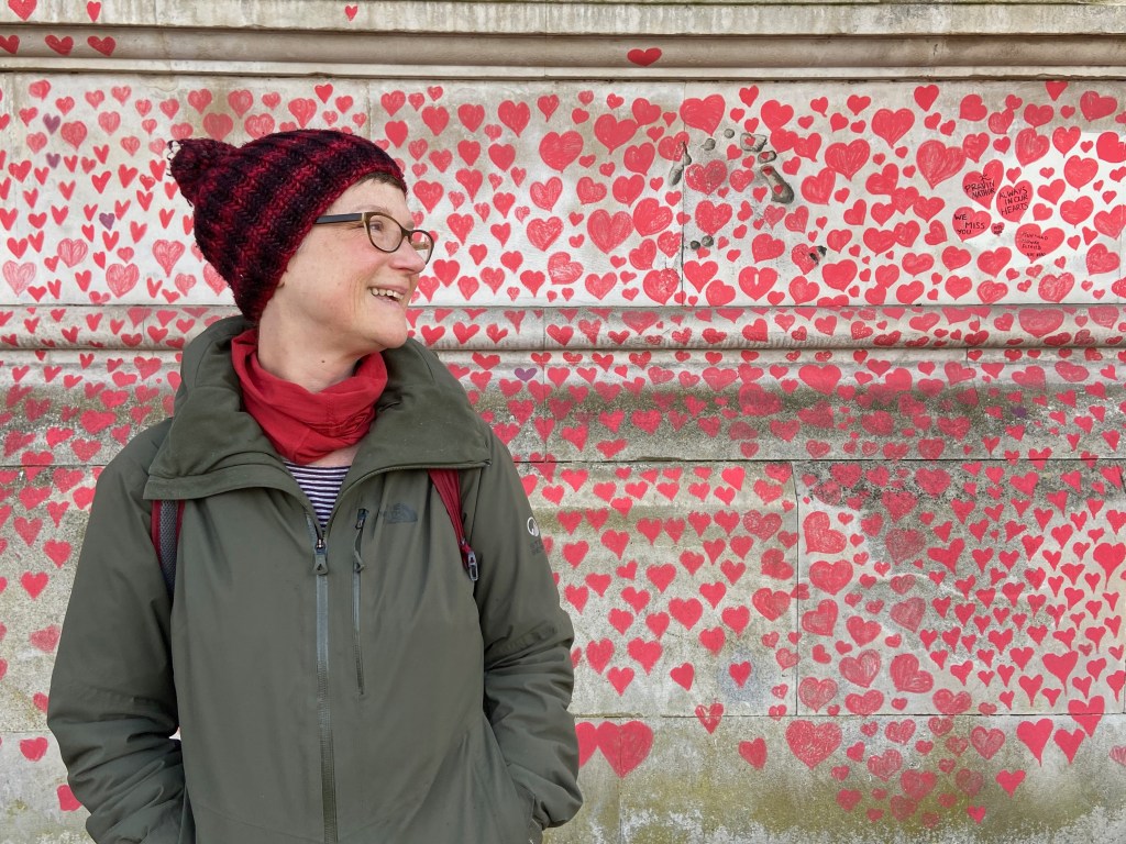 A woman is standing to the left side of the screen. She is bundled up in a warm jacket and winter hat. She is smiling and looking off to the side and there hundreds of hearts behind her.