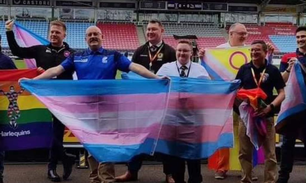 Emmett Peacock holds up a trans Pride flag as other people hold up a variety of LGBTQ+ flags in the background at a rugby pitch