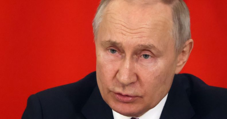 Russia president Vladimir Putin, who has a history of attacking LGBTQ+ people, wears a suit and tie as he sits in front of a red background