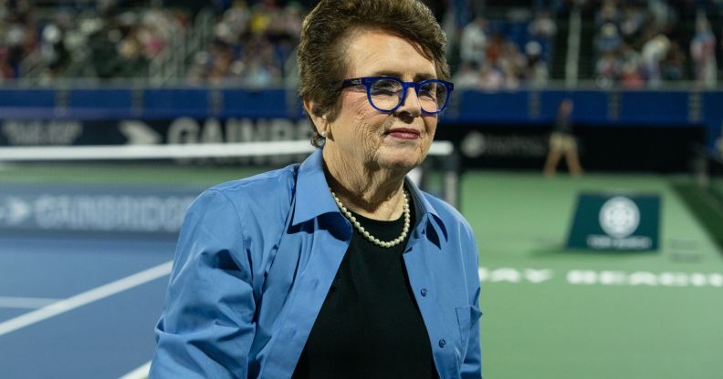 Tennis legend Billie Jean King wears a dark shirt and blue jacket as she stands on a tennis court