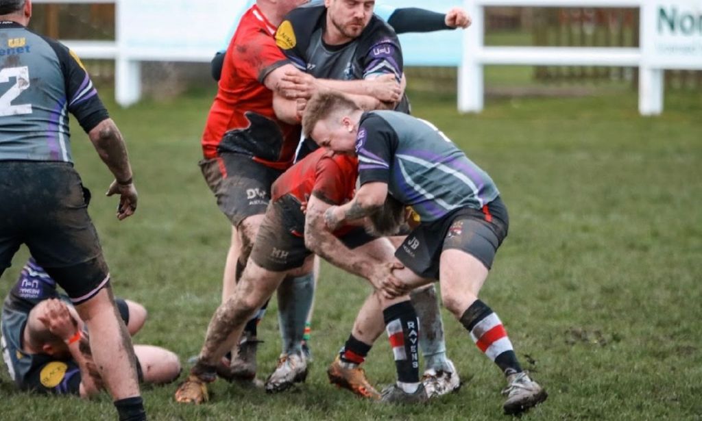 Trans rugby player Jordan Blackwood wears a grey, black and purple rugby uniform as he holds onto a player from a different team during a match