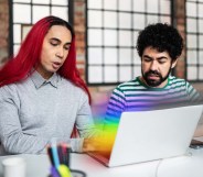 There is a feminine presenting person ont he left side of the photo. They have bright red hair and are wearing a grey shirt. Next to them is a masculine presenting person wearing a beard and a striped shirt. They are both looking down at a laptop computer that is emitting the pride flag colours.