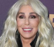 Cher smiling wearing a black coat and blonde hair, stood against a purple background with white writing on it.