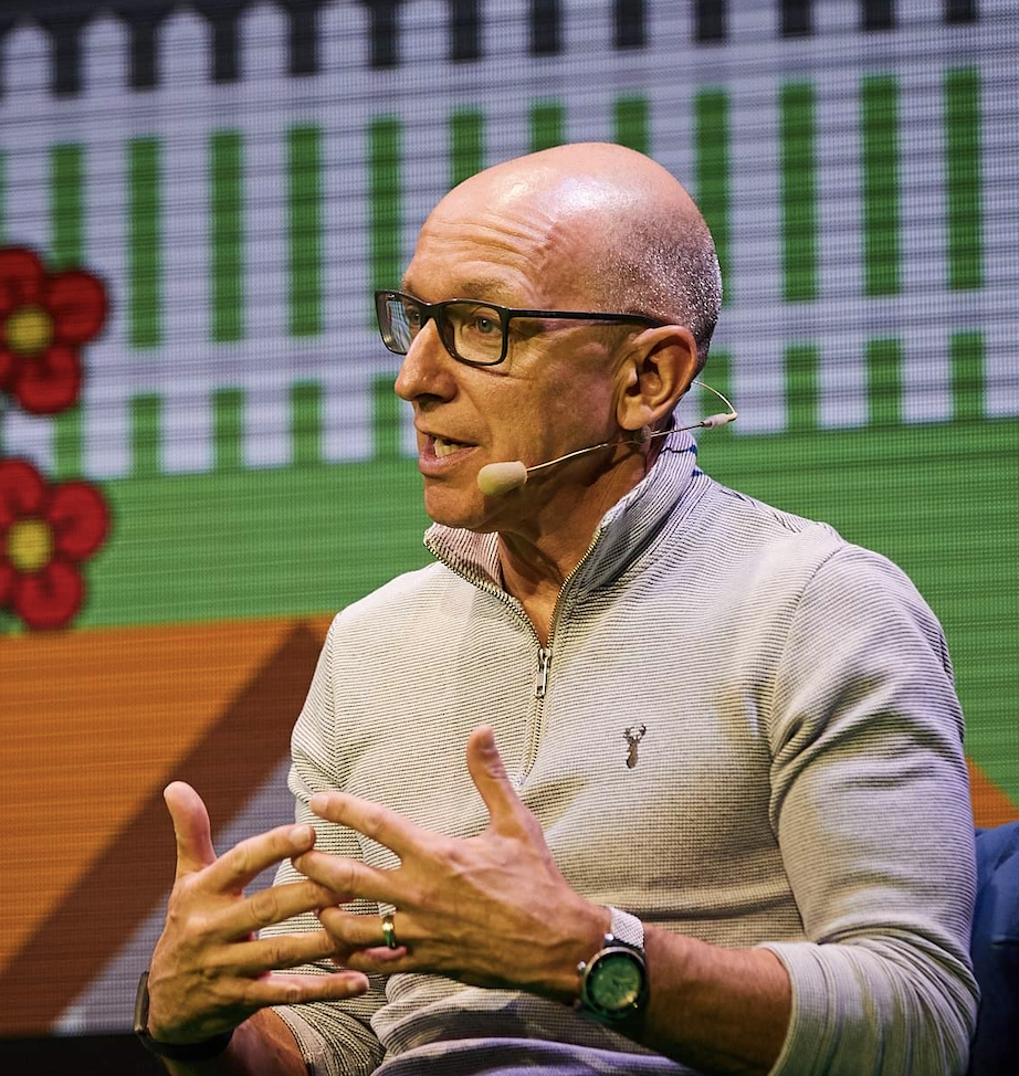 This is an image of Fraser Londgen speaking at a conference. He is bald and is wearing glasses and an off-white cardigan.