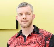 Russell Tovey wearing a black and white shirt while standing in front of a yellow background.