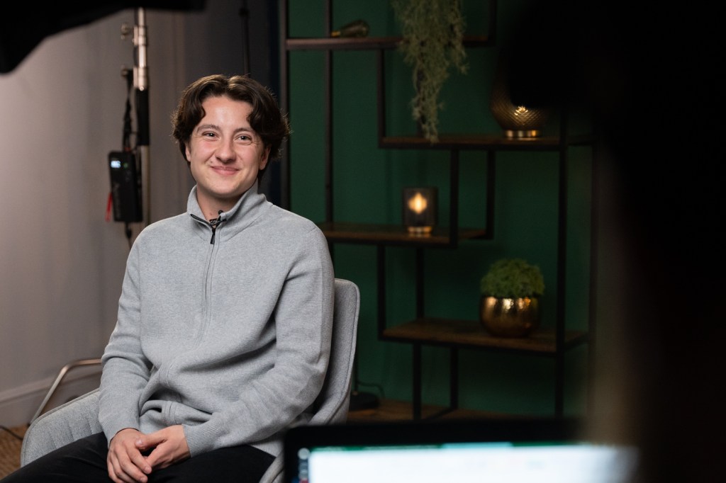 A man is sitting on the left side of the frame. He is wearing a grey cardigan and his hands are folded in his lap. The background is a dark green wall with candles lit.