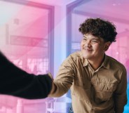In this image, a trans person is wearing a khaki coloured shirt and is shaking hands with someone.