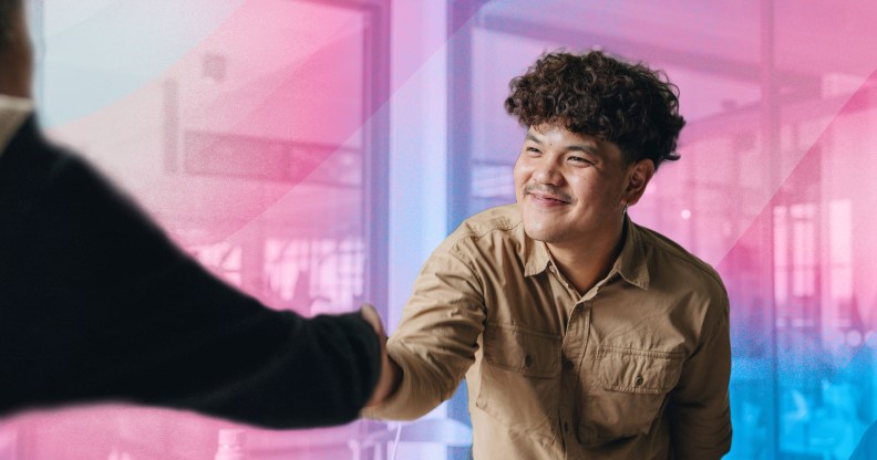 In this image, a trans person is wearing a khaki coloured shirt and is shaking hands with someone.