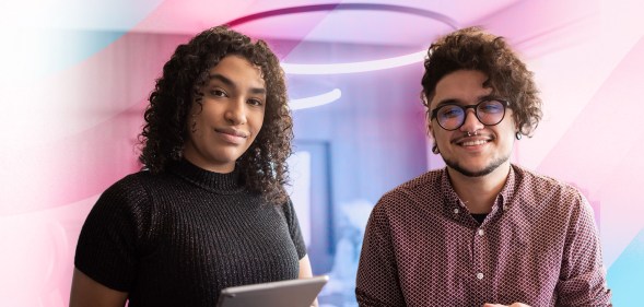 In this image, there are two trans people smiling for the camera. One person is wearing black and holding an iPad and the other is wearing red plaid and glasses.