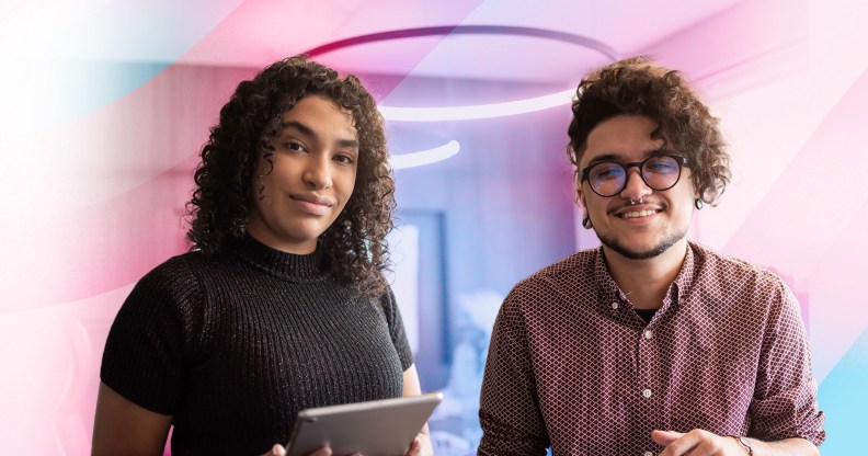 In this image, there are two trans people smiling for the camera. One person is wearing black and holding an iPad and the other is wearing red plaid and glasses.