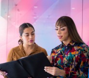 There are two women overlooking a report. One is wearing a brown shirt and the other has a more colourful shirt on. There is a creative background of the trans pride flag colours.