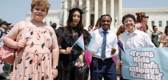 A group of four transgender youth smiling in a photo, with one of them holding a sign that reads "trans kids have always existed."