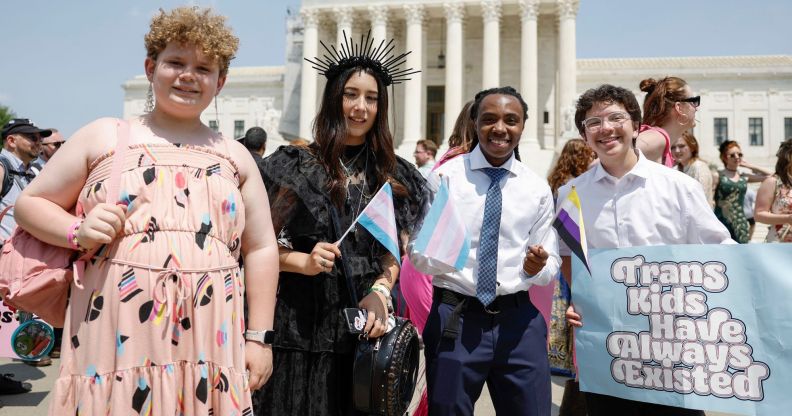 A group of four transgender youth smiling in a photo, with one of them holding a sign that reads "trans kids have always existed."