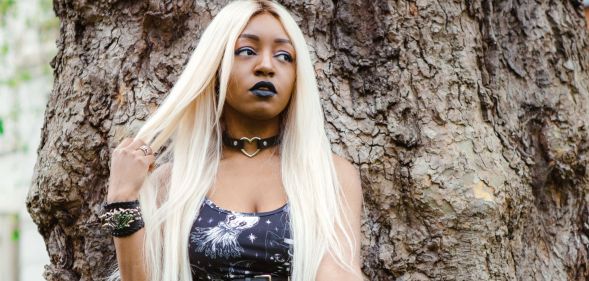 Asexual activist Yasmin Benoit wears a black dress as she poses against a tree and holds part of her blonde hair