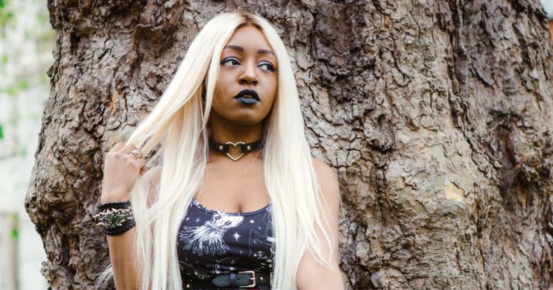 Asexual activist Yasmin Benoit wears a black dress as she poses against a tree and holds part of her blonde hair