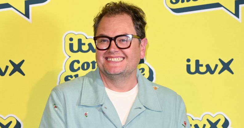 Alan Carr in a mint green shirt and white t-shirt smiling and standing in front of a yellow background.