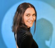 Alyson Stoner wears a black dress and short brown hair while stood against a blue background.