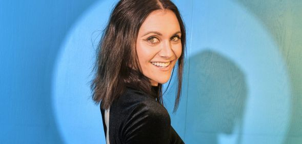 Alyson Stoner wears a black dress and short brown hair while stood against a blue background.