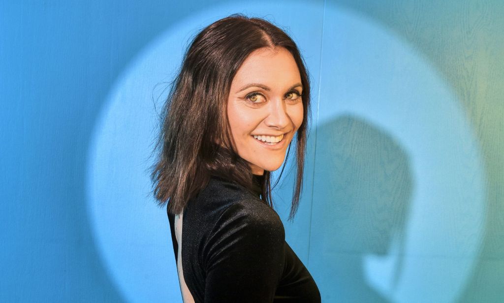 Alyson Stoner wears a black dress and short brown hair while stood against a blue background.