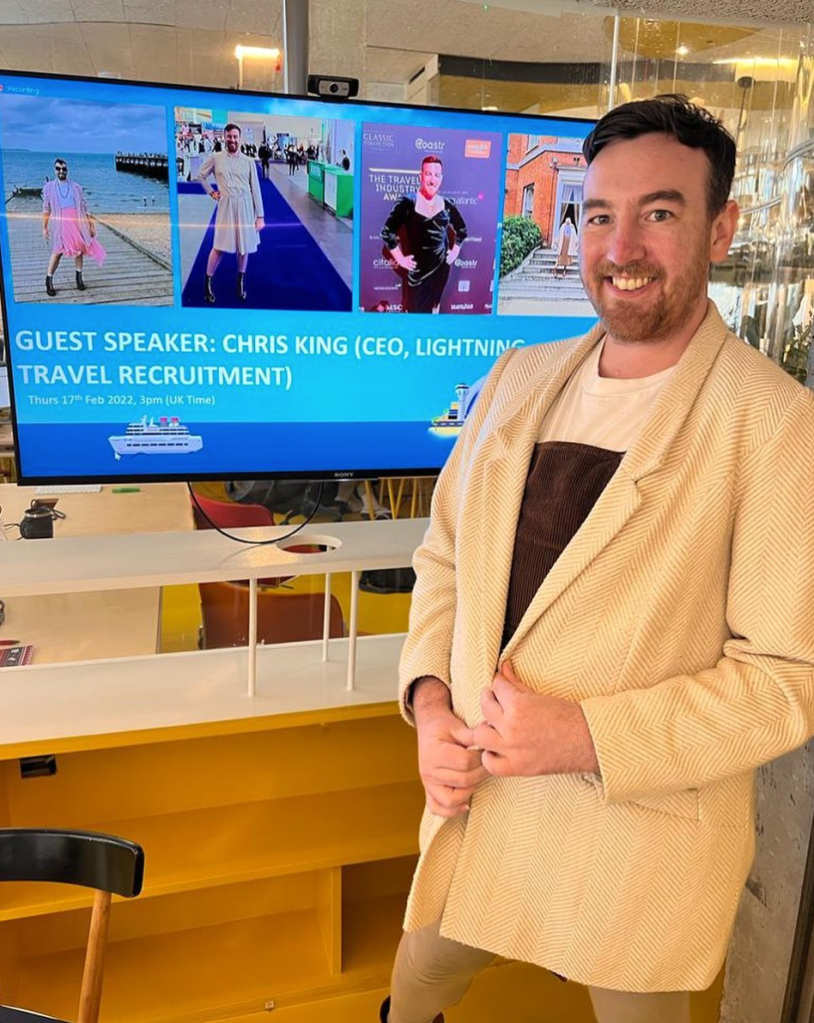 This is a photo of Chris King. They are wearing a tan coloured blazer. They are smiling and standing in front of a monitor.