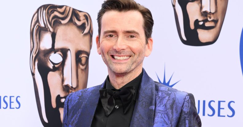 David Tennant wears a blue suit and black shirt while smiling at the camera.