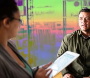 In this image of a job interview, a masculine presenting person wearing green looks on nervously as a woman with glasses is looking down at an iPad.