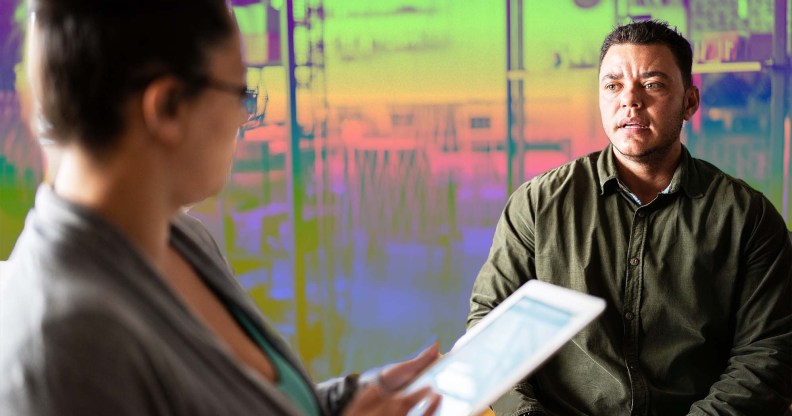 In this image of a job interview, a masculine presenting person wearing green looks on nervously as a woman with glasses is looking down at an iPad.