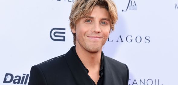 Lukas Gage in a black suit and shirt with parted blonde hair, smiling at the camera.