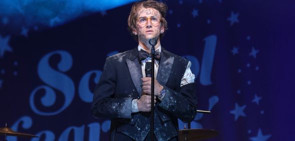 Sebastian Croft wears a filthy suit while standing on a stage and holding a microphone in a new still from new film How To Date Billy Walsh.