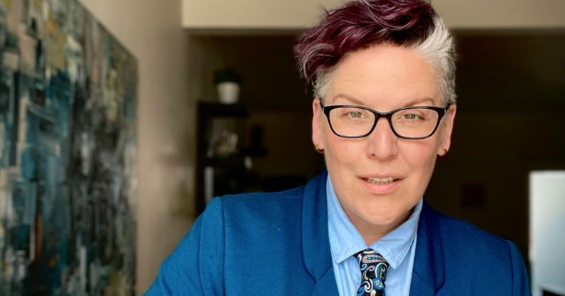 Author and advocate Rowan Jetté Knox wears a blue shirt, dark blue tie with a paisley pattern and medium toned blue jacket as they pose for the camera during their tour around the UK to talk about trans rights and the power of allyship