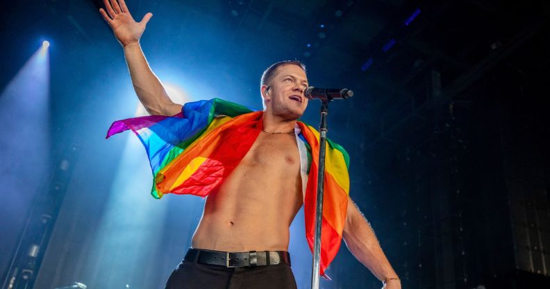 Imagine Dragons frontman Dan Reynolds performs shirtless on stage with an LGBTQ+ flag wrapped around his shoulders.