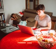 In this image, a woman is sitting at a table and working on her computer. There is a red tablecloth. There are two kids in the background.