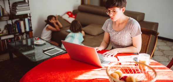 In this image, a woman is sitting at a table and working on her computer. There is a red tablecloth. There are two kids in the background.