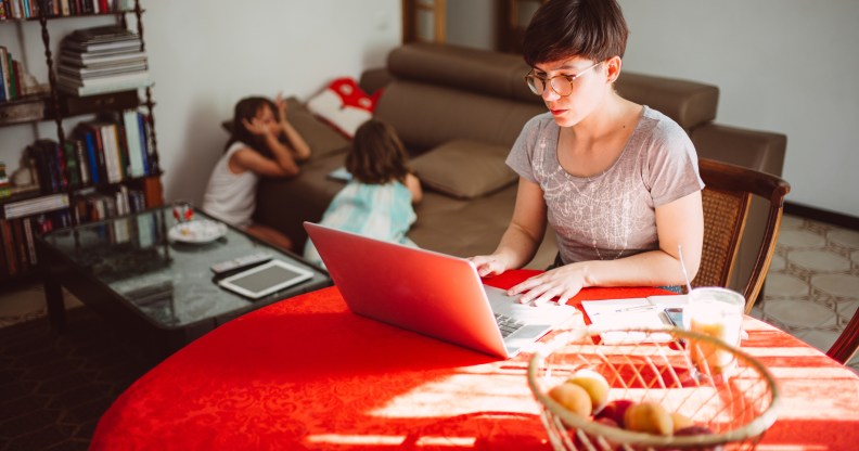 In this image, a woman is sitting at a table and working on her computer. There is a red tablecloth. There are two kids in the background.