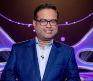 Paul Sinha in a blue suit and shirt standing in front of a blue and purple background.