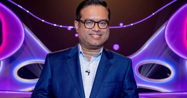 Paul Sinha in a blue suit and shirt standing in front of a blue and purple background.