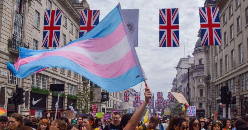 A person holds a trans flag in the air at a crowded protest, with four union jacks being hung across two buildings.