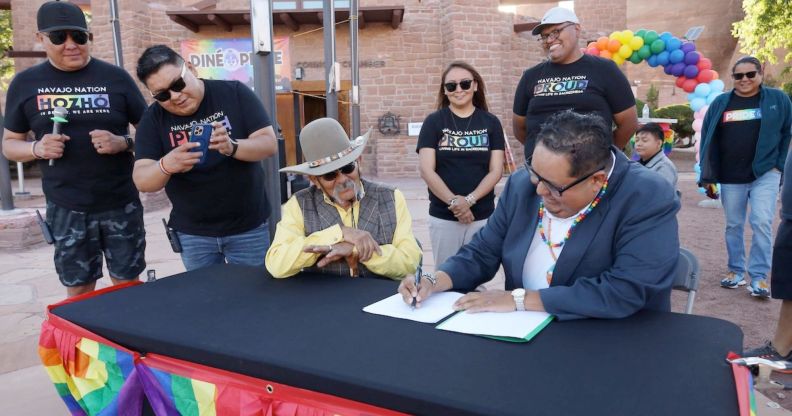 An audience dressed in Pride t shirts gather around Seth Damon as he signs legislation on a desk covered with Pride flags.