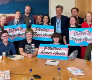 A group of MPs and trans youth sit around a wooden table, holding signs that say "protect trans youth" and "listen to trans youth."