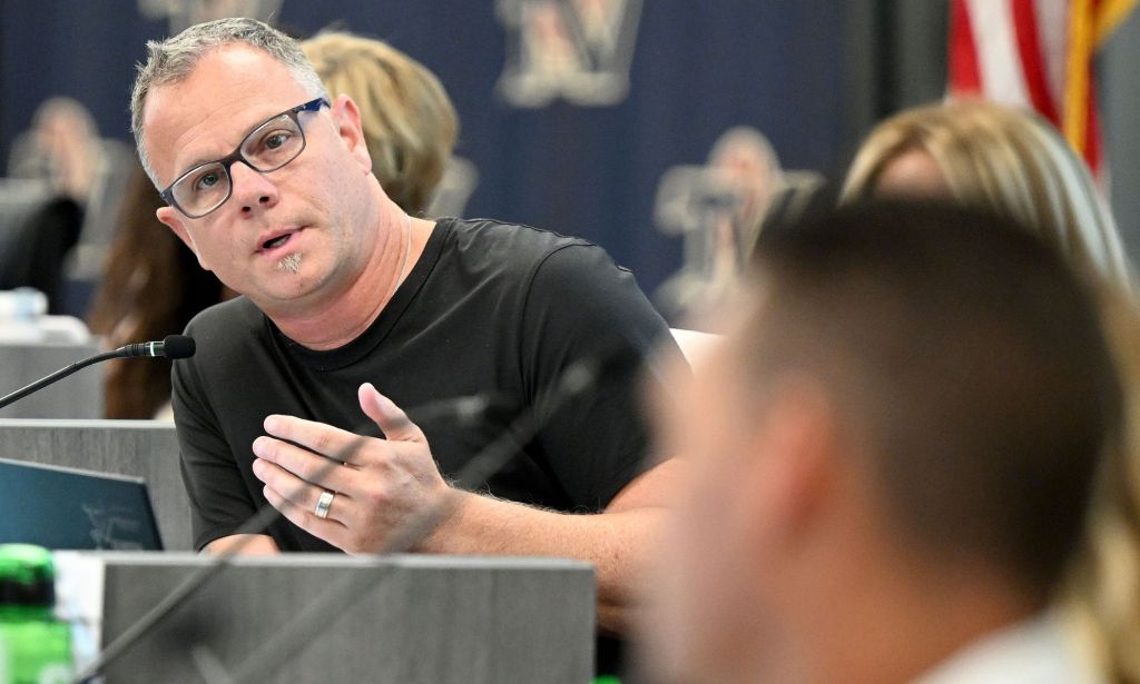 Temecula Valley Unified School District president Dr Joseph Komrosky  wears a black shirt as he speaks into a microphone while a meeting takes place to discuss materials mentioning Harvey Milk