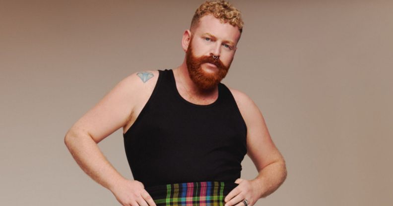 Non-binary, trans singer Tom Rasmussen wears a black vest and tartan shorts against a beige background.