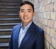 This is an image of Wayne Ting, the ceo of Lime. He is leaning on a brick wall. He is wearing a dark blue blazer with a light blue shirt He has black hair.