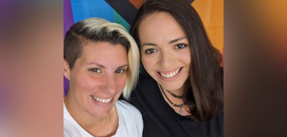 LGBTQ+ foster carers Nat and Terri stand side-by-side as they smile for the camera in front of a Progressive Pride flag