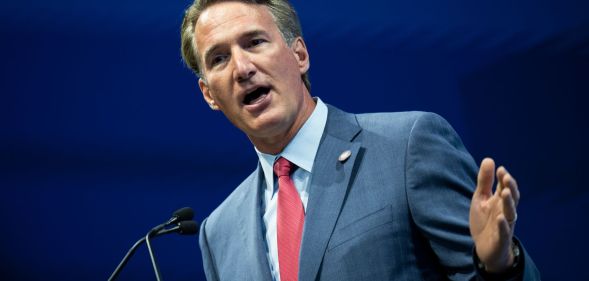 Virginia governor Glenn Youngkin wears a blue shirt, red tie and blue suit jacket as he gestures with one hand raised while standing at a podium