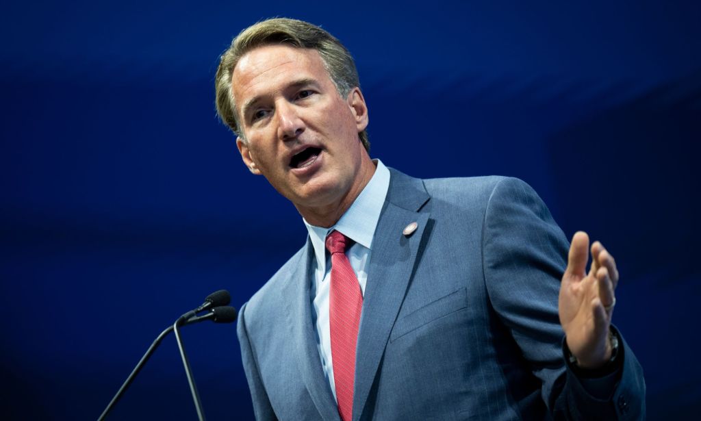 Virginia governor Glenn Youngkin wears a blue shirt, red tie and blue suit jacket as he gestures with one hand raised while standing at a podium