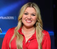 Kelly Clarkson smiling during a radio appearance. She is wearing a red shirt and two necklaces.