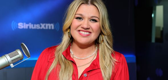 Kelly Clarkson smiling during a radio appearance. She is wearing a red shirt and two necklaces.