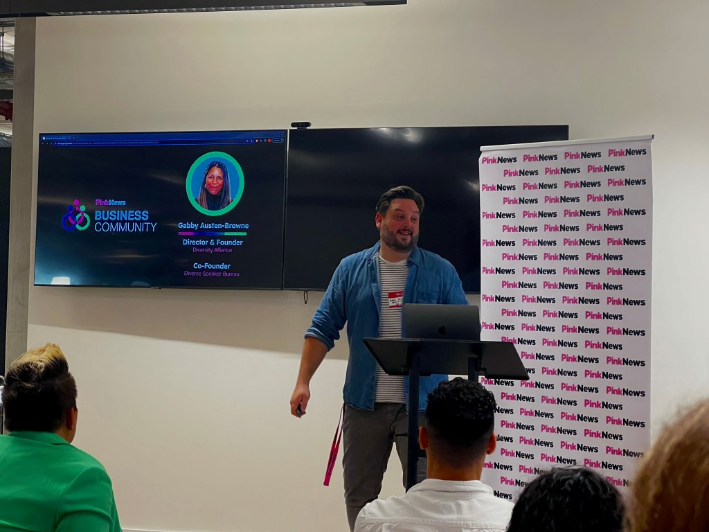 This is an image of Mark Shields speaking at an event. He is wearing khaki trousers with a denim shirt. He is in front of branding that says "Pinknews" 