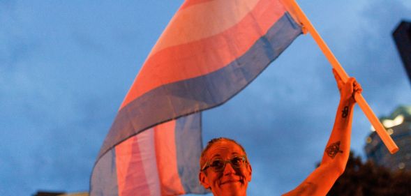 A person holds up a trans flag, both of which are bathed in an orange glow.